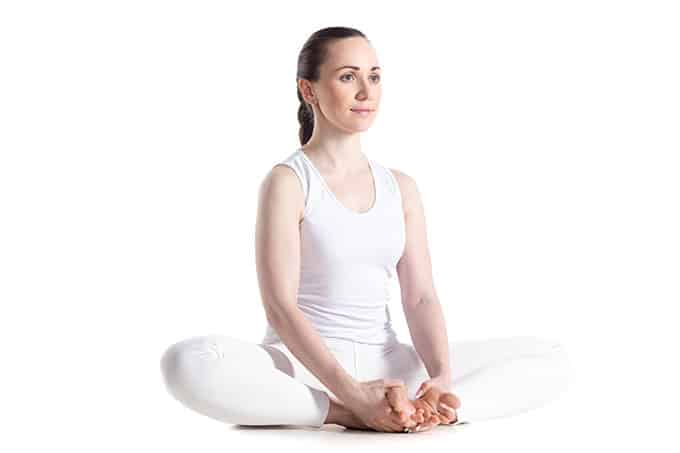 A woman wearing white sitting on the ground and holding her feet together in front of her as she stretches her legs outwards so her knees touch the ground